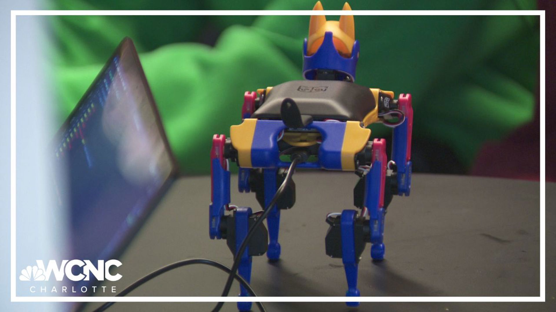 A colorful quadruped robot beside a laptop, with the WCNC Charlotte logo